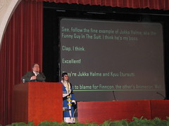 Jukkahoo behind the podium, Kyuu Eturautti standing beside it; funny translations on the screen in the back