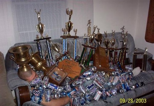 drunk guy buried underneath hordes of beer cans and 15 or more trophies (possibly won in beer drinking contests?)